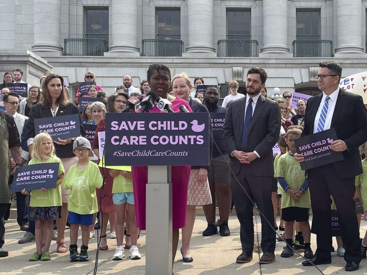 A speaker speaks at a podium that says "Save Child Care Counts" and is surrounded by people, and the state Capitol in the background.